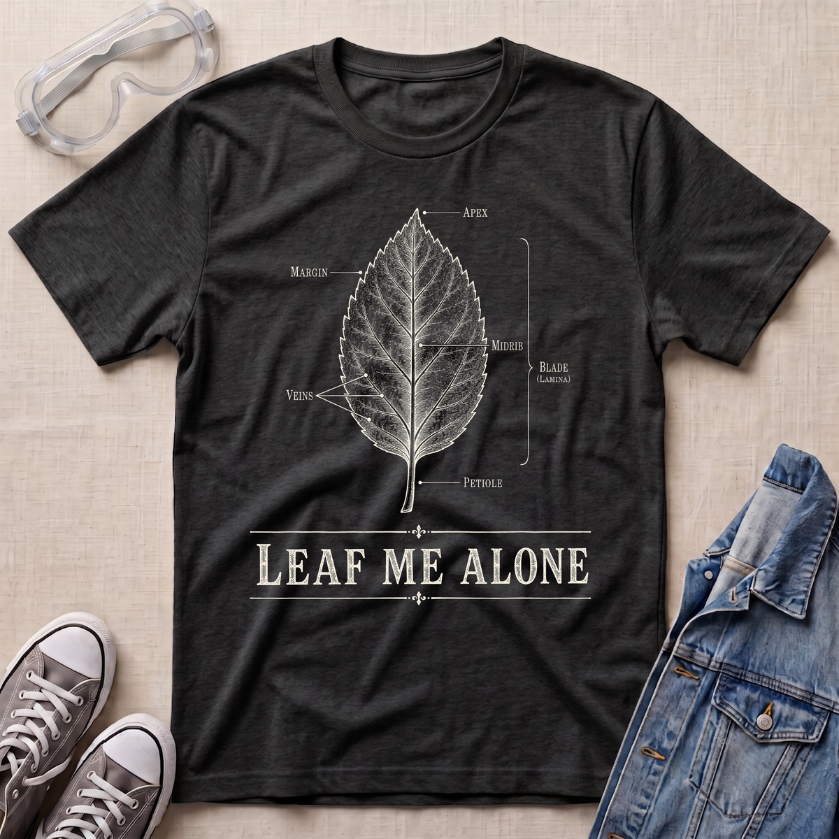 A dark heather grey botanical t-shirt featuring a vintage labeled leaf anatomy illustration with the pun “Leaf Me Alone” in bold retro typography.