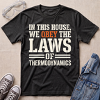 A dark heather gray graphic t-shirt laid flat on a beige fabric background, styled with safety goggles, gray sneakers, and a denim jacket. The t-shirt features the text 'IN THIS HOUSE, WE OBey THE LAWS OF THERMODYNAMICS' in white and orange lettering.