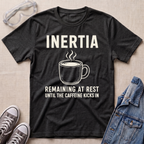 A dark heather gray graphic t-shirt laid flat on a beige textured background, surrounded by lifestyle props including safety goggles, gray sneakers, and a denim jacket. The t-shirt features a white graphic of a steaming coffee mug with the text 'INERTIA REMAINING AT REST UNTIL THE CAFFEINE KICKS IN'.