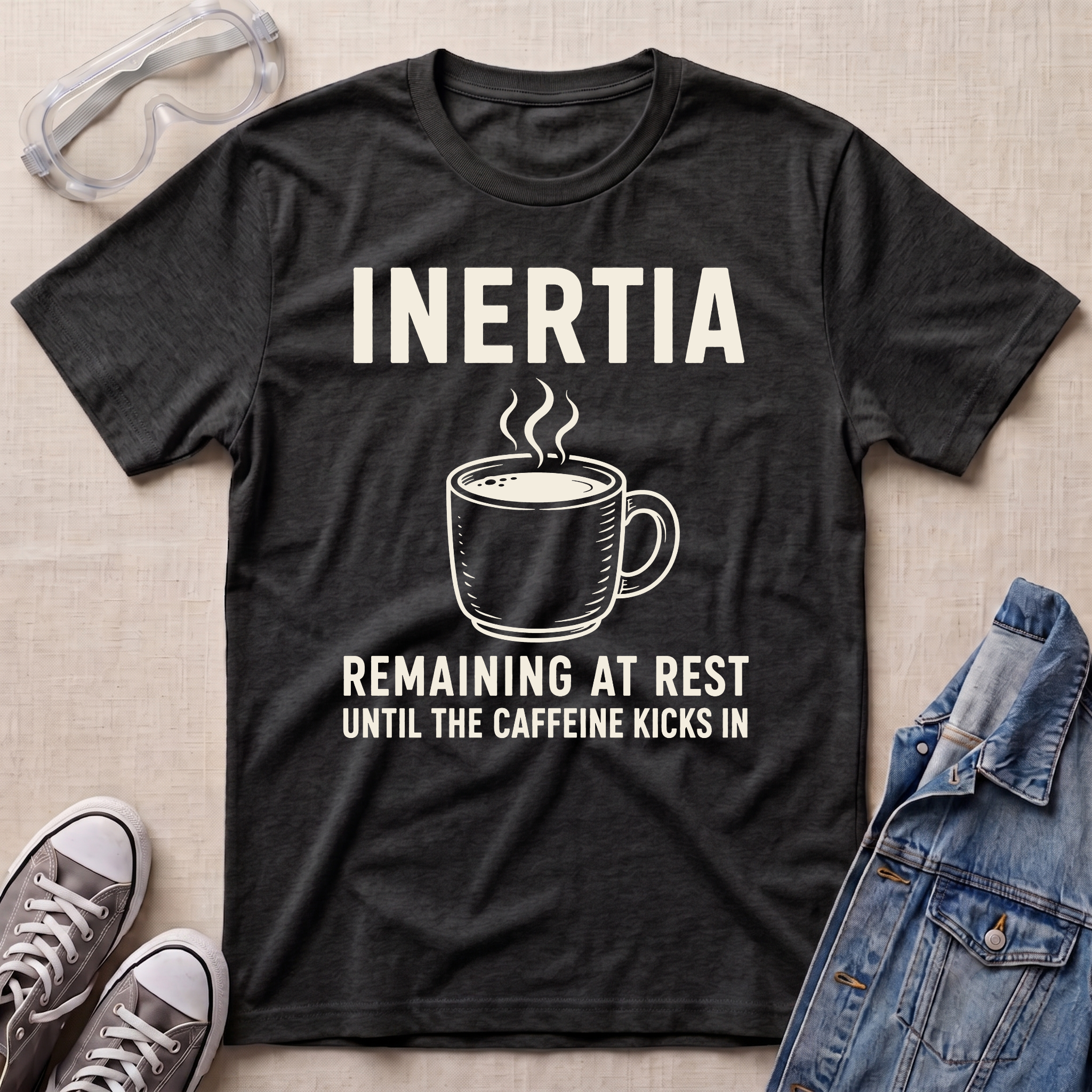 A dark heather gray graphic t-shirt laid flat on a beige textured background, surrounded by lifestyle props including safety goggles, gray sneakers, and a denim jacket. The t-shirt features a white graphic of a steaming coffee mug with the text 'INERTIA REMAINING AT REST UNTIL THE CAFFEINE KICKS IN'.