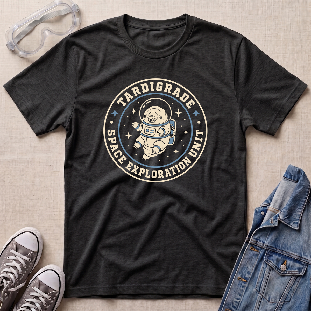 A dark heather grey t-shirt laid flat on a light beige textured background, styled with safety goggles, gray sneakers, and a denim jacket. The t-shirt features a circular graphic of an astronaut floating in space with the text 'TARDIGRADE SPACE EXPLORATION UNIT'.