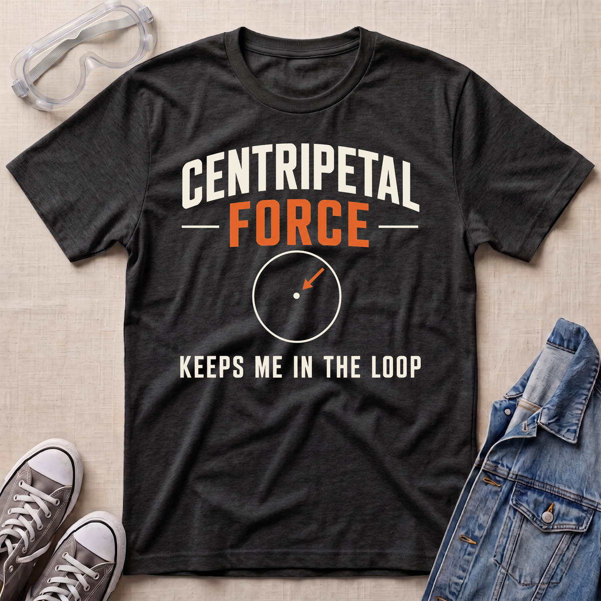 A dark heather gray graphic t-shirt laid flat on a light beige textured background, styled with safety goggles, gray sneakers, and a blue denim jacket. The t-shirt features the text 'CENTRIPETAL FORCE' in white and orange, with a small compass graphic below.