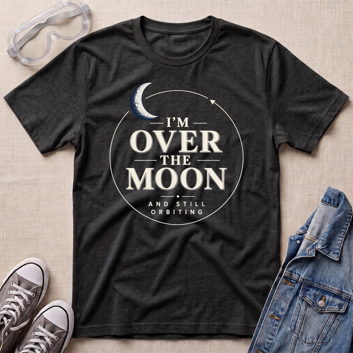 A dark heather grey graphic t-shirt laid flat on a beige fabric background, styled with safety goggles, gray sneakers, and a denim jacket. The t-shirt features a white circular graphic with a crescent moon and the text 'I'M OVER THE MOON AND STILL ORBITING'.