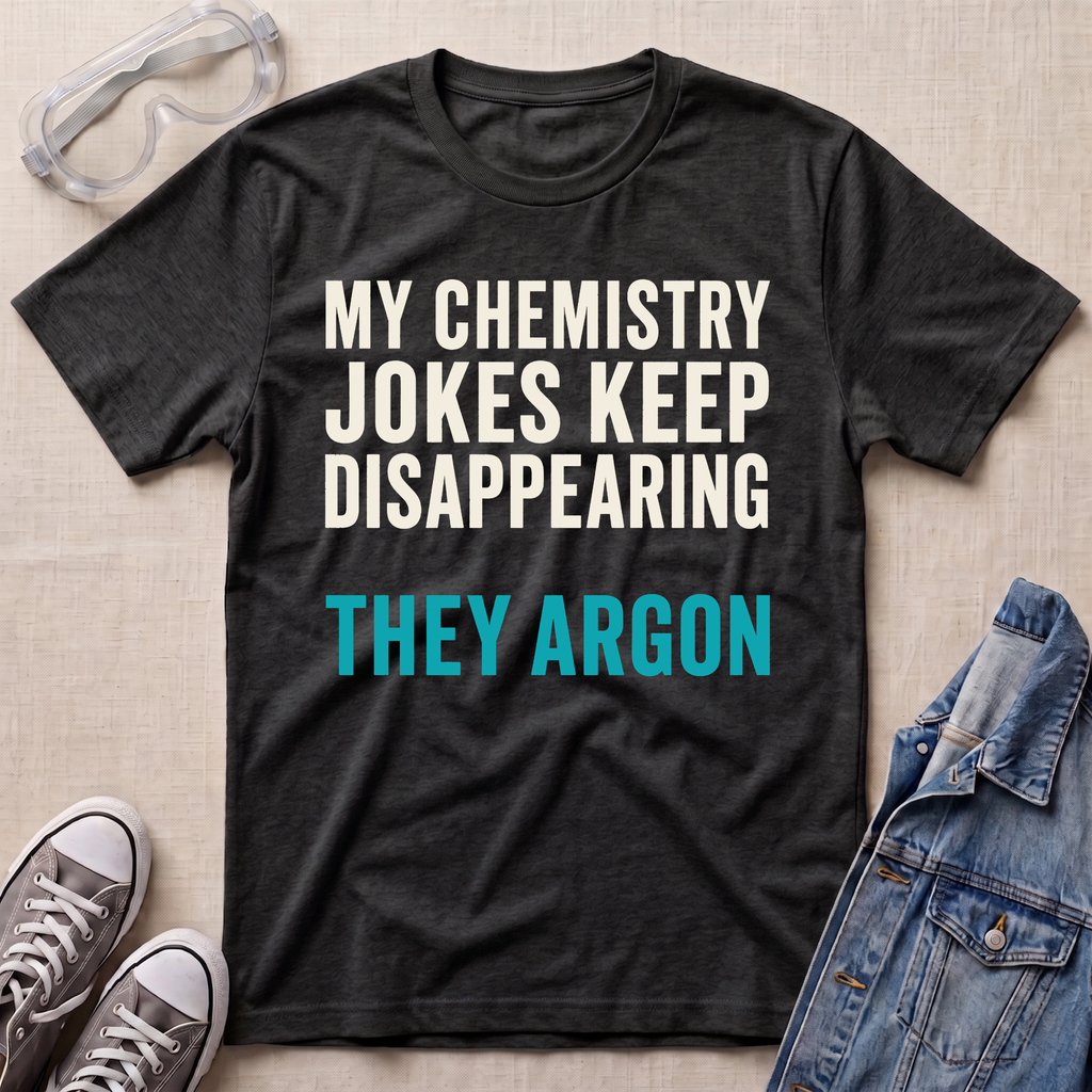A dark heather gray graphic t-shirt laid flat on a beige textured background, surrounded by styling props including safety goggles, gray sneakers, and a blue denim jacket. The t-shirt features the text 'MY CHEMISTRY JOKES KEEP DISAPPEARING' in white and 'THEY ARGON' in bright teal blue.