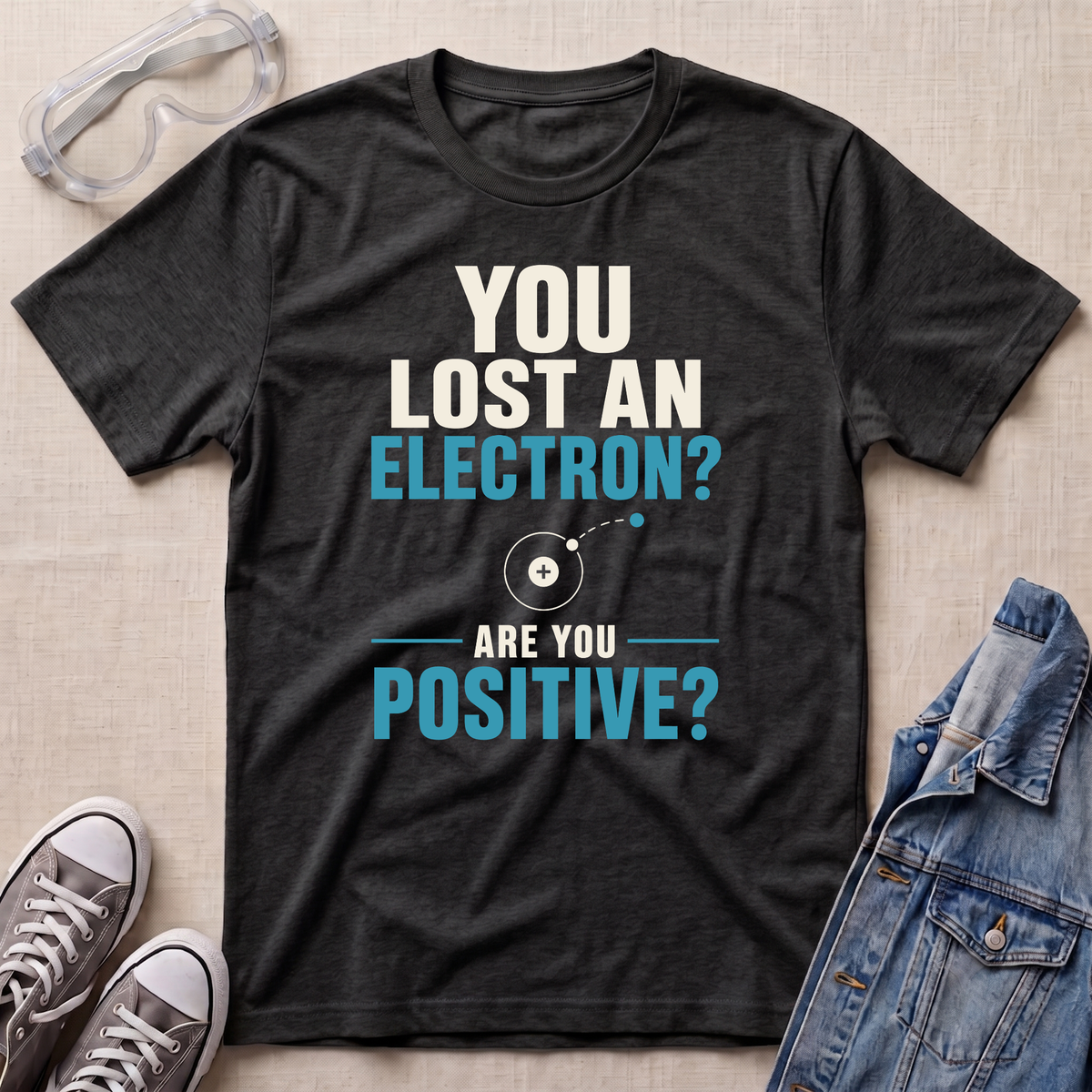 A dark heather gray graphic t-shirt laid flat on a beige textured background, surrounded by lifestyle props including safety goggles, gray sneakers, and a denim jacket. The t-shirt features a white and light blue text design that reads 'YOU LOST AN ELECTRON? ARE YOU POSITIVE?' with a simple atom diagram.