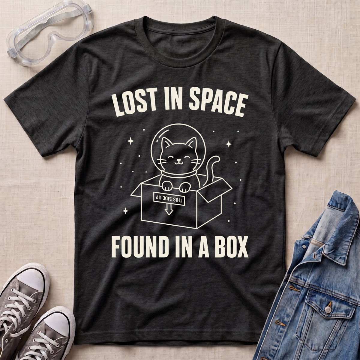 A dark heather gray graphic t-shirt laid flat on a beige fabric background, surrounded by styling props including clear safety glasses, gray sneakers, and a blue denim jacket. The t-shirt features a white graphic of a cat inside a box with a helmet and the text 'LOST IN SPACE FOUND IN A BOX'.