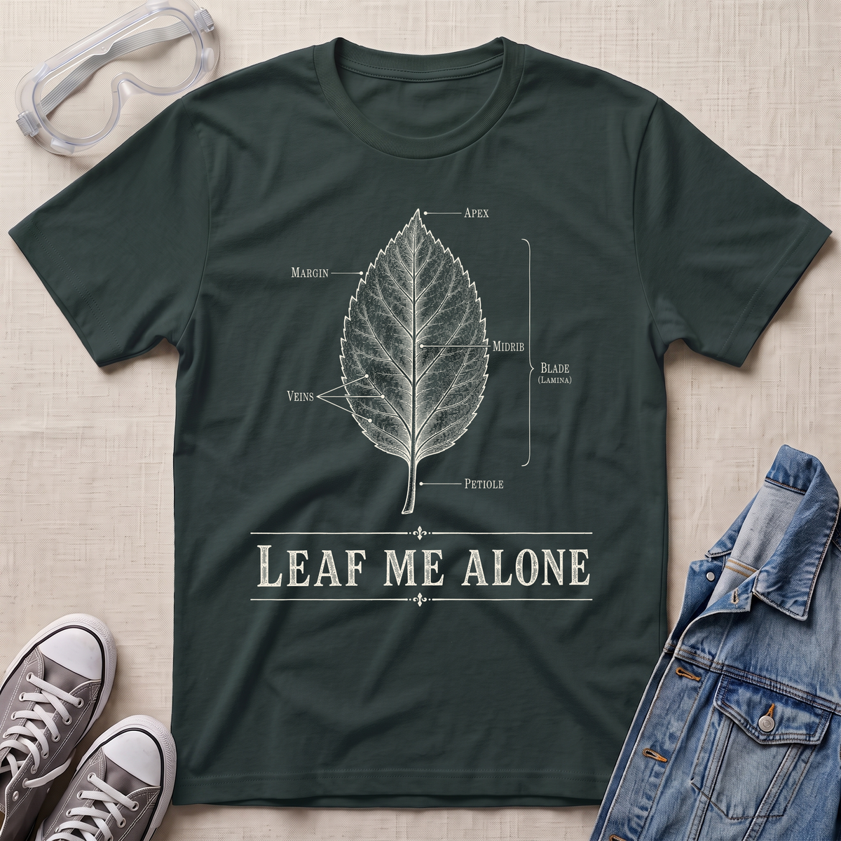 A dark forest green botanical t-shirt featuring a vintage labeled leaf anatomy illustration with the pun “Leaf Me Alone” in bold retro typography.
