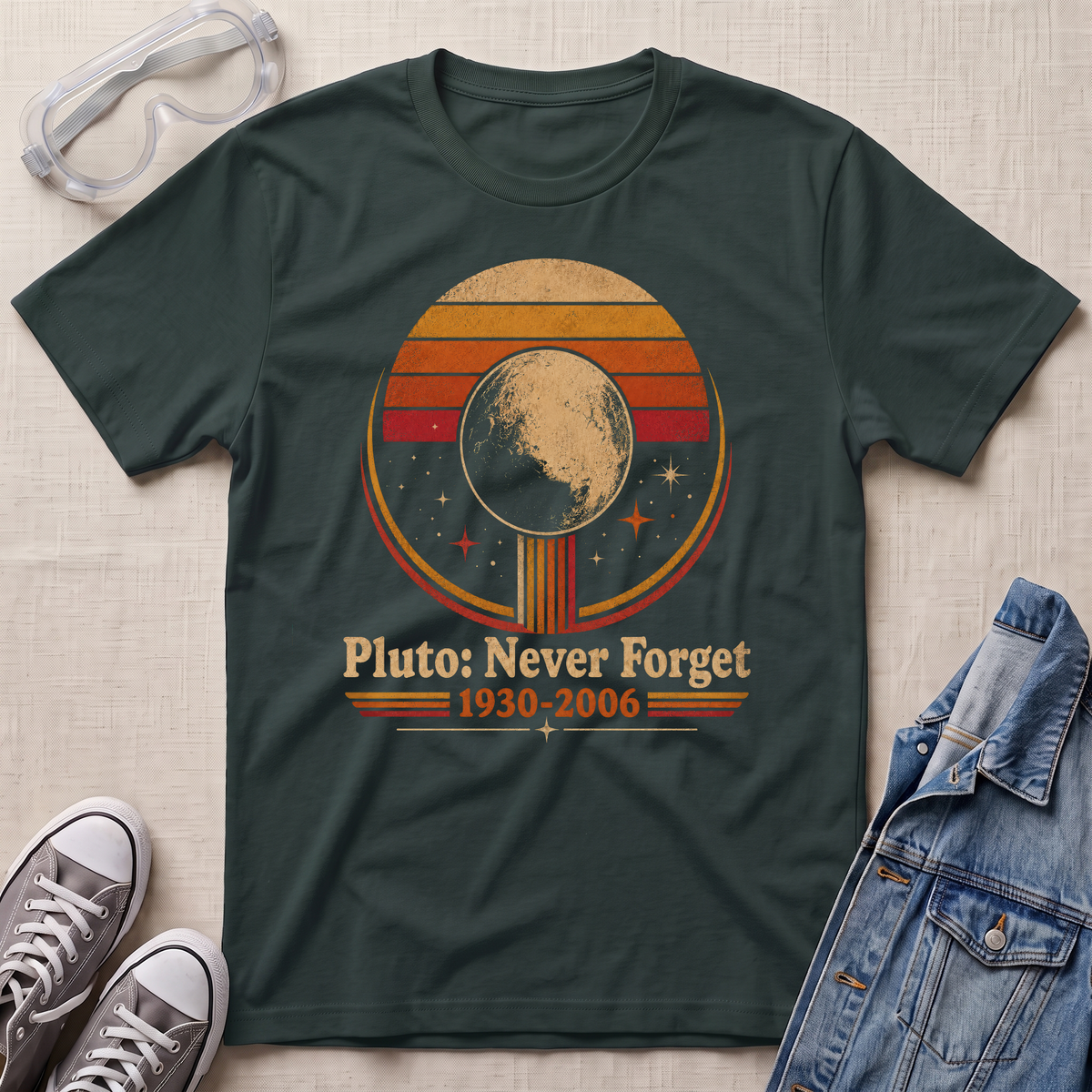 A dark green graphic t-shirt laid flat on a light beige fabric background, styled with safety goggles, gray sneakers, and a denim jacket. The t-shirt features a retro circular graphic of Pluto with the text 'Pluto: Never Forget 1930-2006'.