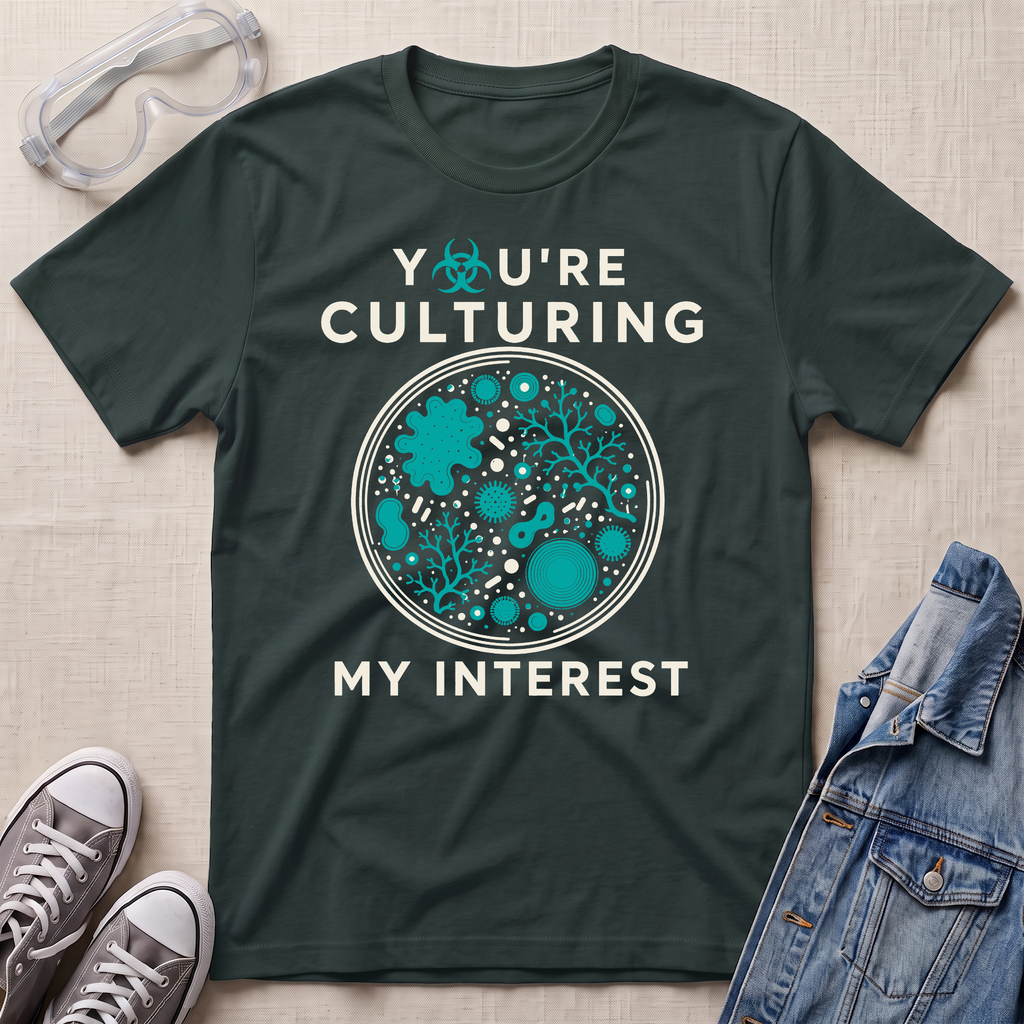 A dark green graphic t-shirt laid flat on a beige fabric background, styled with safety goggles, gray sneakers, and a denim jacket. The t-shirt features a circular petri-dish style graphic in teal and white with the text 'YOU'RE CULTURING MY INTEREST' and a small biohazard symbol.