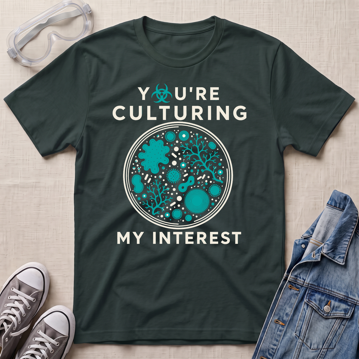 A dark green graphic t-shirt laid flat on a beige fabric background, styled with safety goggles, gray sneakers, and a denim jacket. The t-shirt features a circular petri-dish style graphic in teal and white with the text 'YOU'RE CULTURING MY INTEREST' and a small biohazard symbol.