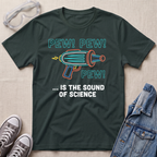 A dark green graphic t-shirt laid flat on a light beige fabric background, styled with safety goggles, gray sneakers, and a denim jacket. The t-shirt features a colorful retro toy gun illustration with the text 'PEW! PEW! PEW! ... IS THE SOUND OF SCIENCE.'