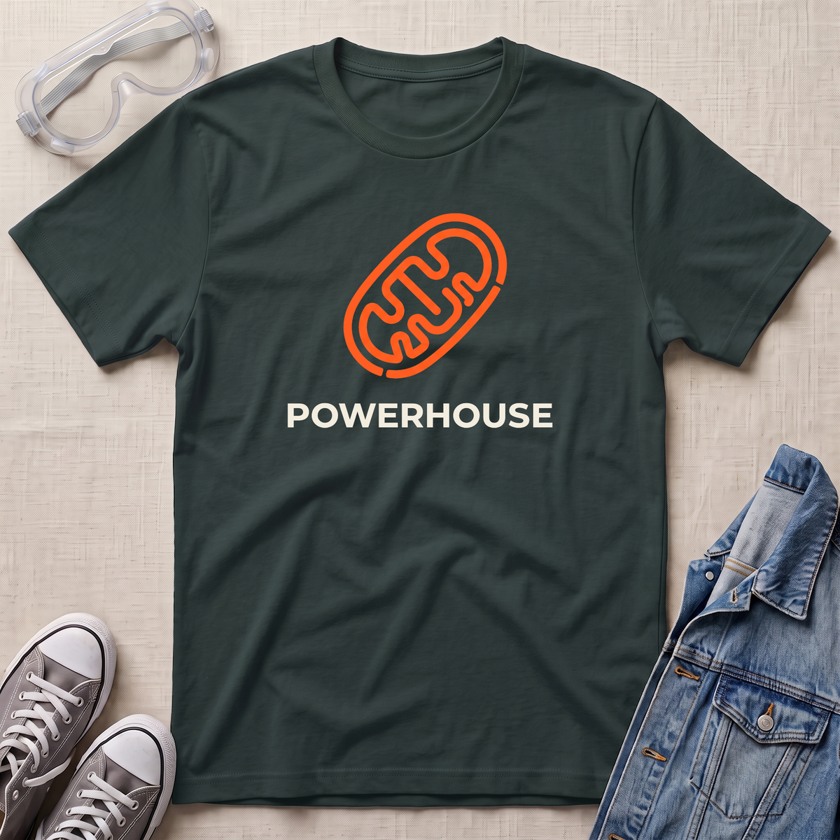 A dark green crewneck t-shirt with an orange mitochondrial outline and the word 'POWERHOUSE' in white, styled in a flat lay with safety goggles, sneakers, and a denim jacket on a beige fabric background.