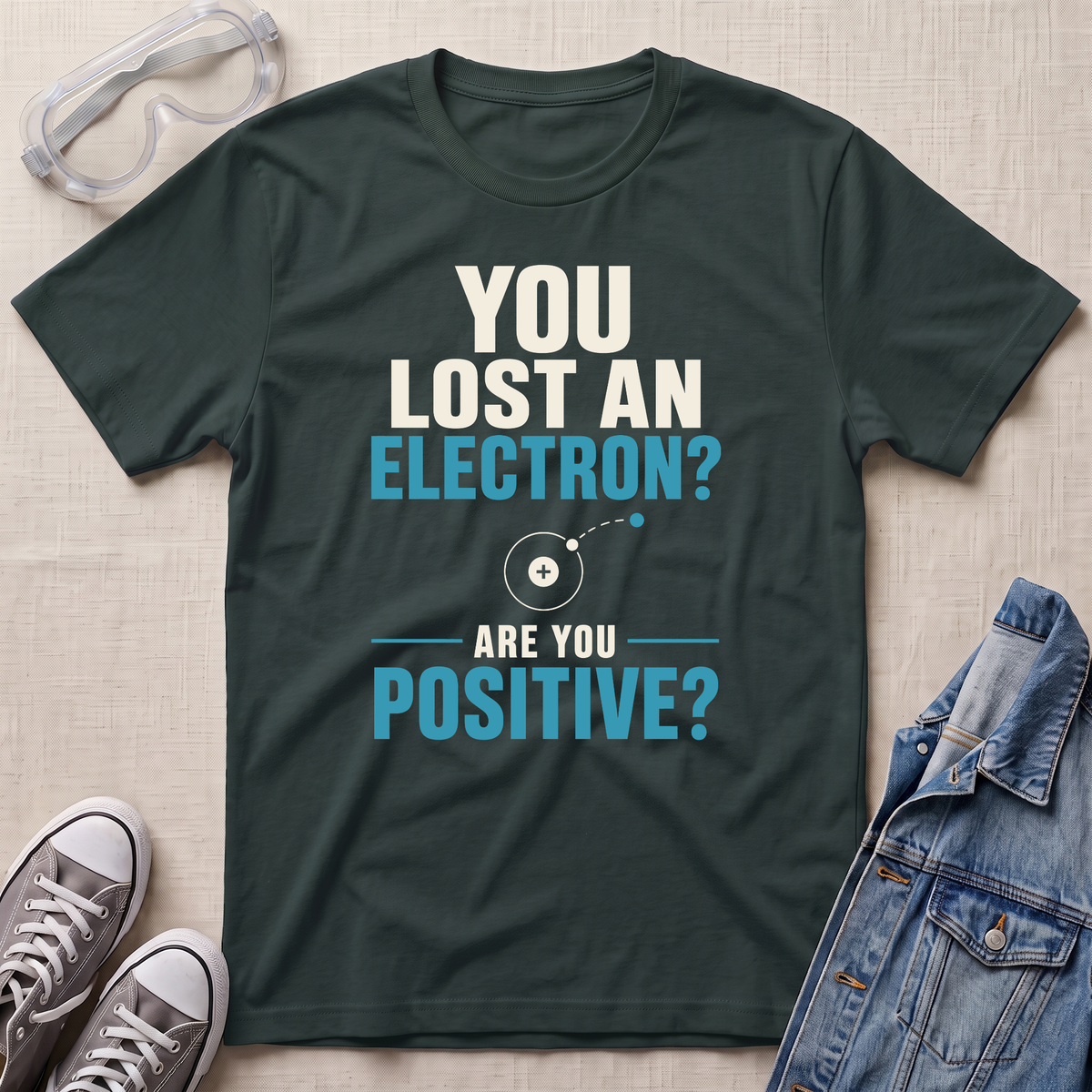 A dark green graphic t-shirt laid flat on a light beige textured surface, styled with safety goggles, gray sneakers, and a denim jacket. The t-shirt features a white and light blue text design that reads 'YOU LOST AN ELECTRON? ARE YOU POSITIVE?' with a simple atom diagram.