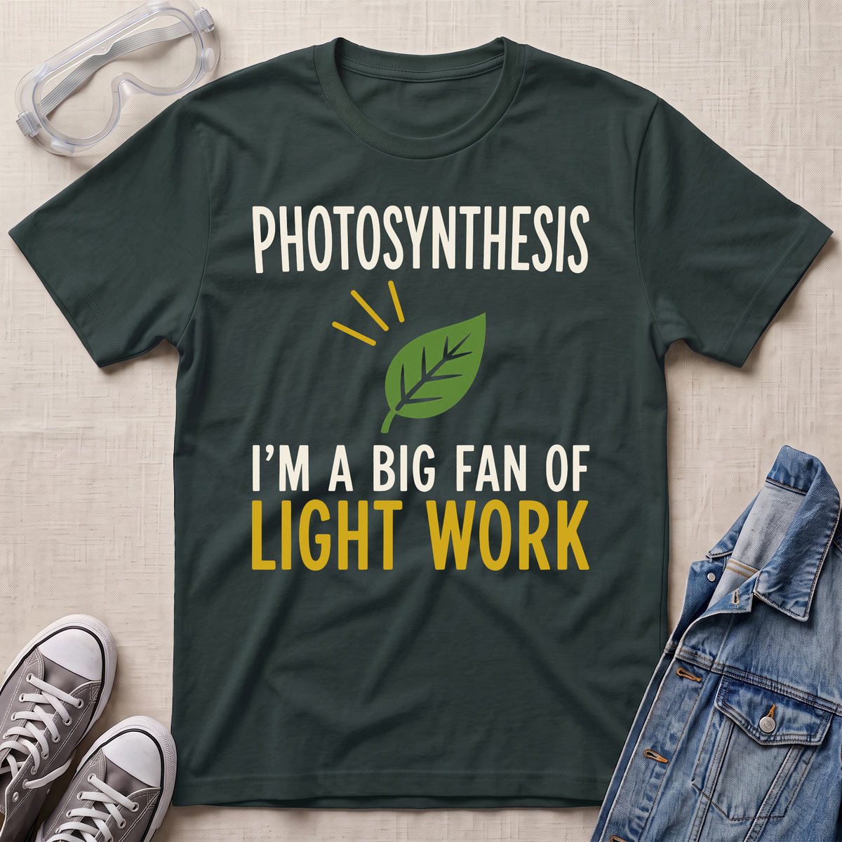 A dark green graphic t-shirt laid flat on a beige fabric background, styled with safety goggles, gray sneakers, and a denim jacket. The t-shirt features a white and yellow text graphic that reads 'PHOTOSYNTHESIS I'M A BIG FAN OF LIGHT WORK' with a green leaf illustration.