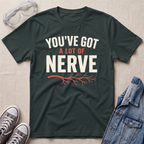 A dark green graphic t-shirt laid flat on a light beige textured background, surrounded by styling props including safety goggles, gray sneakers, and a denim jacket. The t-shirt features the text 'YOU'VE GOT A LOT OF NERVE' in white and orange lettering with an orange branch graphic underneath.