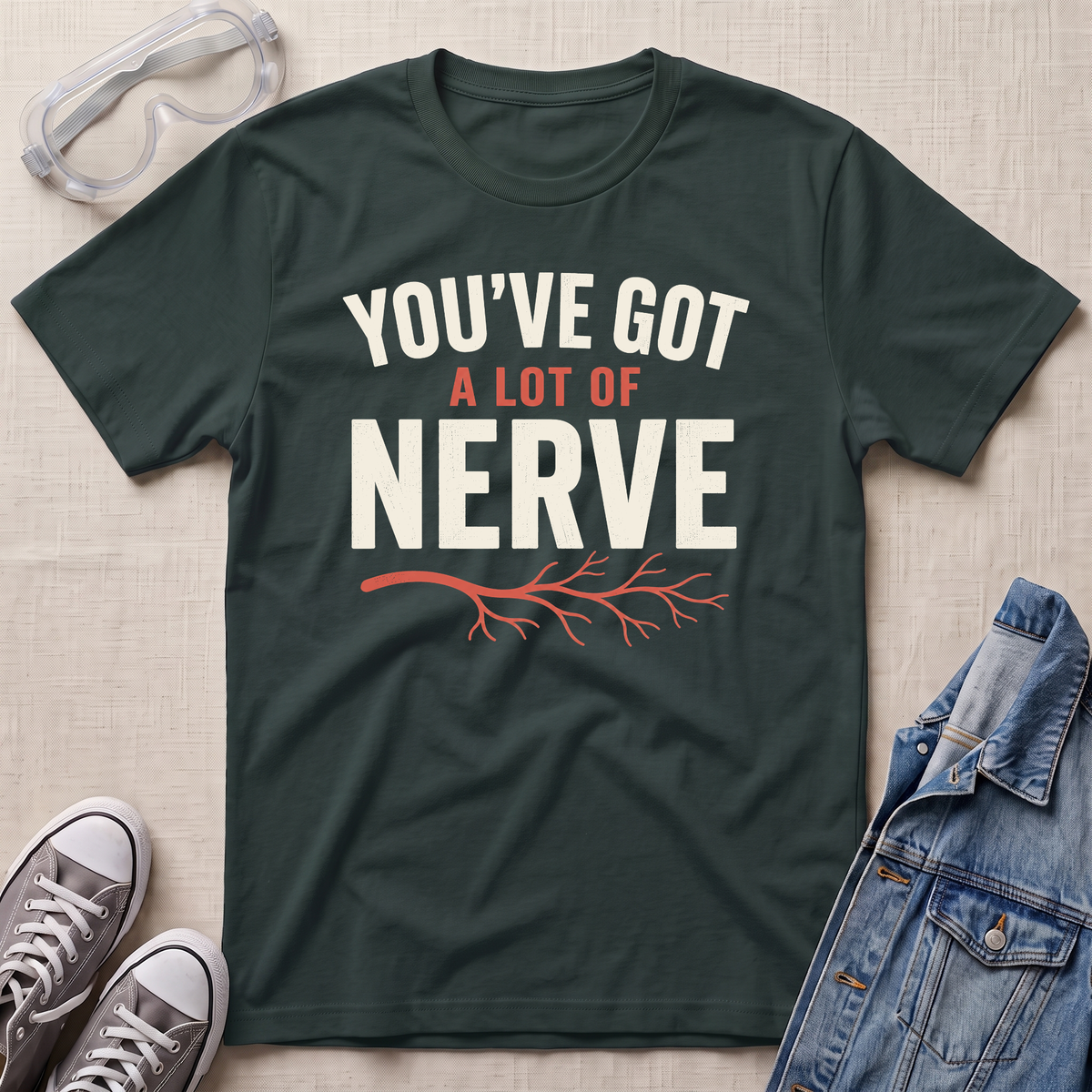 A dark green graphic t-shirt laid flat on a light beige textured background, surrounded by styling props including safety goggles, gray sneakers, and a denim jacket. The t-shirt features the text 'YOU'VE GOT A LOT OF NERVE' in white and orange lettering with an orange branch graphic underneath.