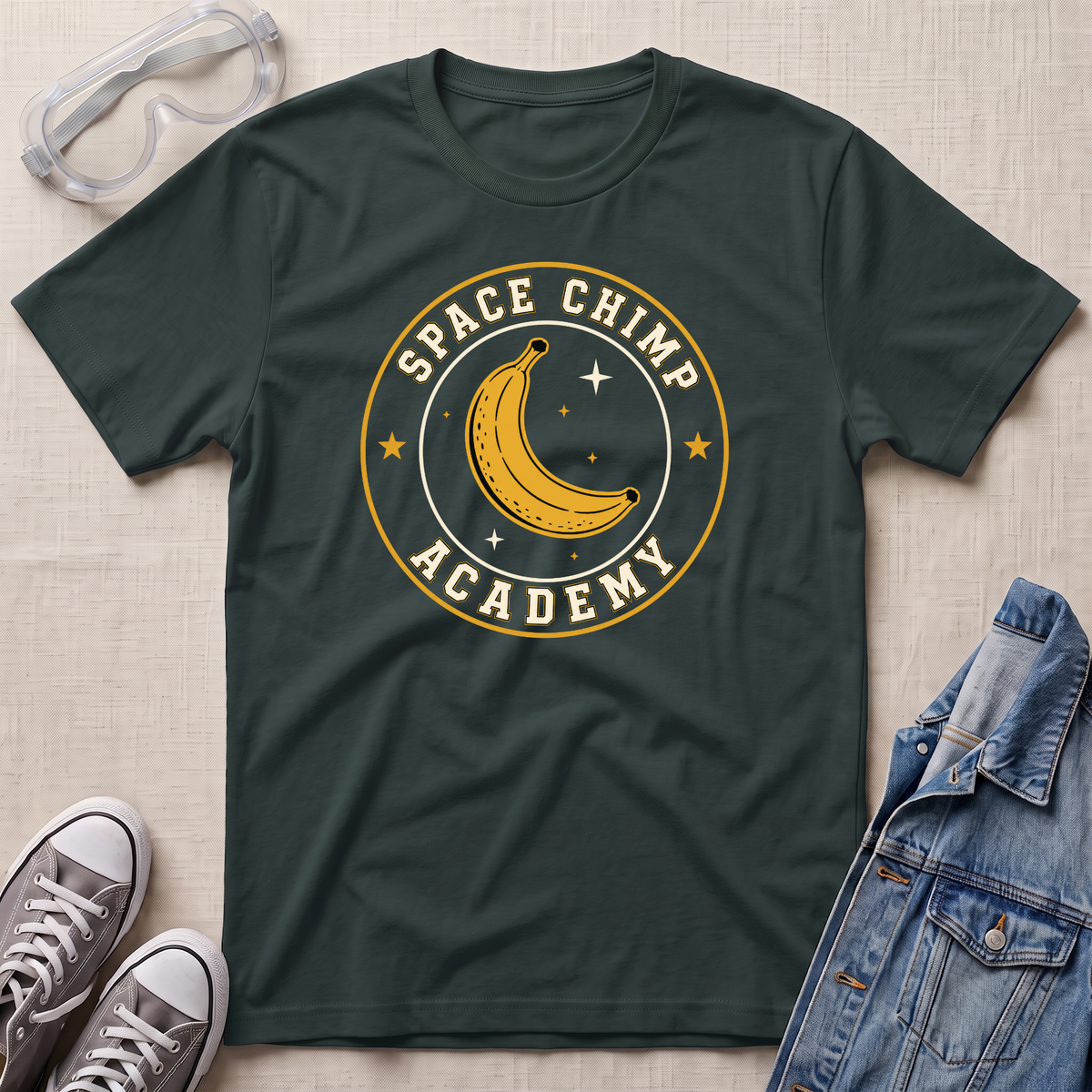 A dark forest green crewneck t-shirt with a 'Space Chimp Academy' graphic featuring a yellow banana and stars, styled in a flat lay with safety goggles, gray sneakers, and a denim jacket on a beige fabric background.