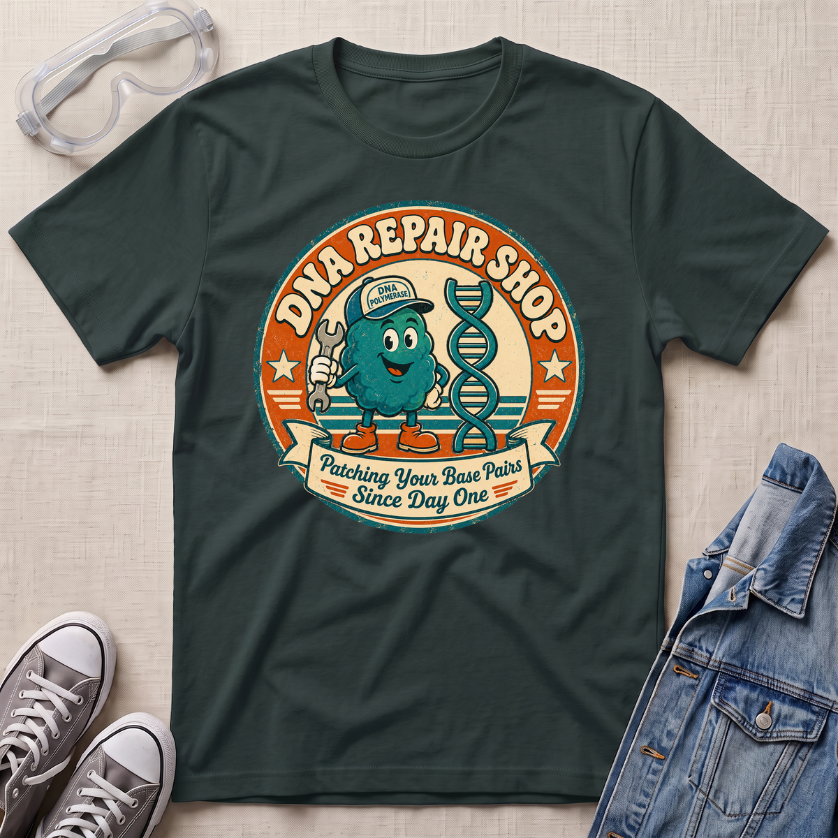 A dark green graphic t-shirt laid flat on a light beige textured surface, surrounded by styling props including clear safety glasses, gray sneakers, and a blue denim jacket. The t-shirt features a retro circular graphic for 'DNA REPAIR SHOP' with a cartoon teal DNA helix character and the slogan 'Patching Your Base Pairs Since Day One'.