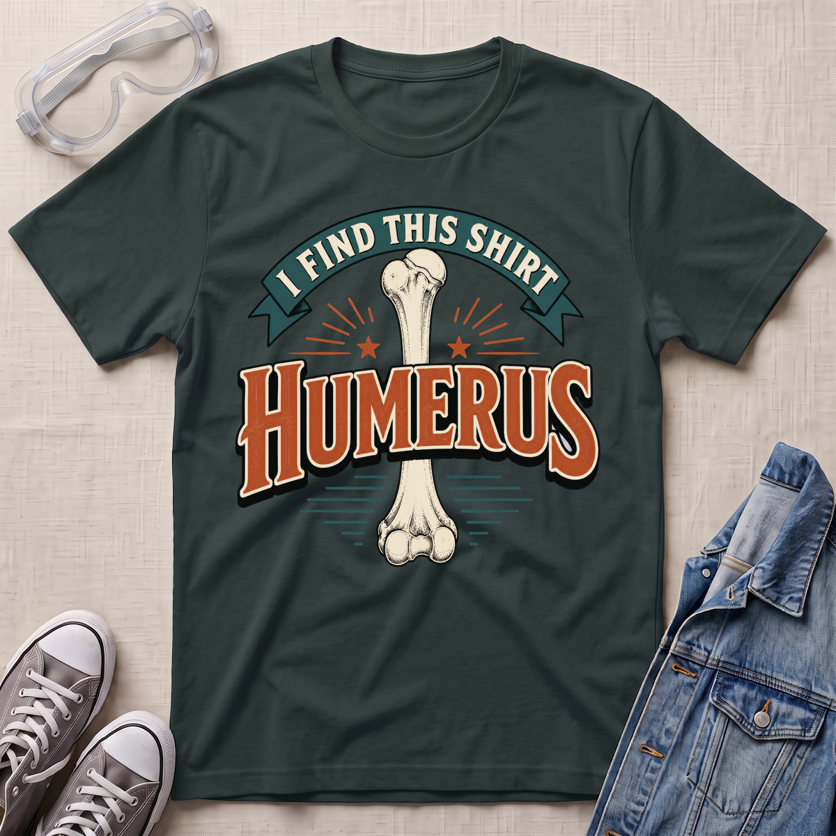 A dark forest green graphic t-shirt laid flat on a light wood surface, featuring a bone graphic and the text 'I FIND THIS SHIRT HUMERUS' in orange and green. The shirt is styled with safety glasses, gray sneakers, and a denim jacket.