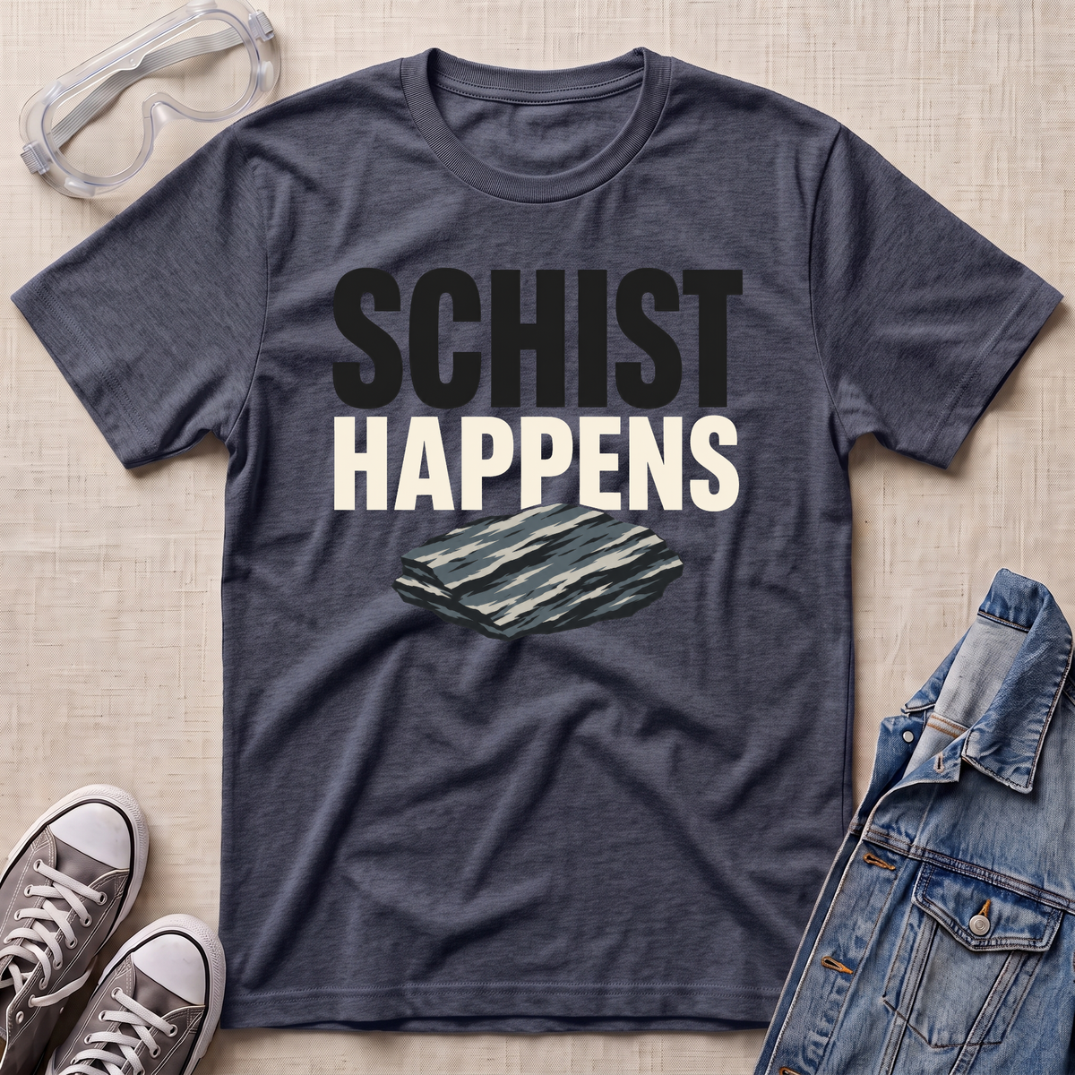 A heather navy graphic t-shirt laid flat on a light beige textured background, surrounded by styling props including safety glasses, gray sneakers, and a denim jacket. The t-shirt features the text 'SCHIST HAPPENS' in bold black and white lettering above an illustration of a layered schist rock.