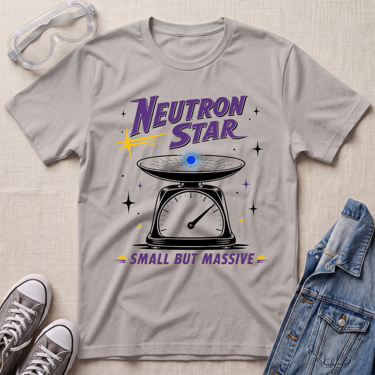A light ice gray graphic t-shirt laid flat on a textured beige surface, styled with safety goggles, gray sneakers, and a denim jacket. The t-shirt features a retro sci-fi graphic of a weighing scale with a glowing blue sphere and purple text reading 'Neutron Star' and 'Small But Massive'.
