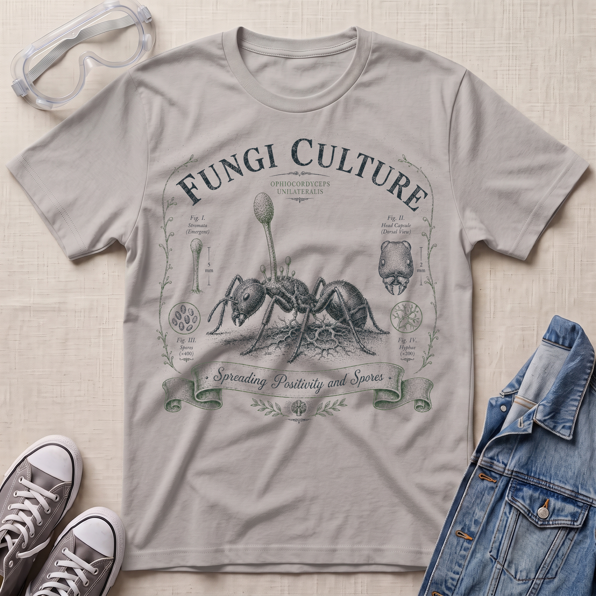 A light gray graphic T-shirt laid flat on a light fabric surface, surrounded by styling props including clear safety glasses, gray sneakers, and a blue denim jacket. The shirt features a vintage-style printed design reading “Fungi Culture” with an ant illustration and decorative text.