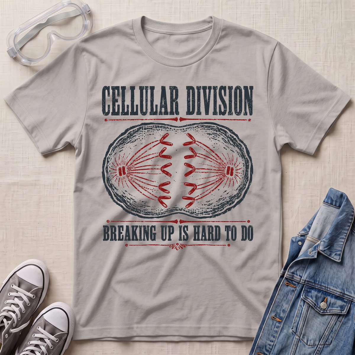 A light gray graphic t-shirt laid flat on a textured beige fabric background, styled with safety glasses, gray sneakers, and a blue denim jacket. The t-shirt features a distressed print of a cell division diagram with the text 'CELLULAR DIVISION' and 'BREAKING UP IS HARD TO DO'.
