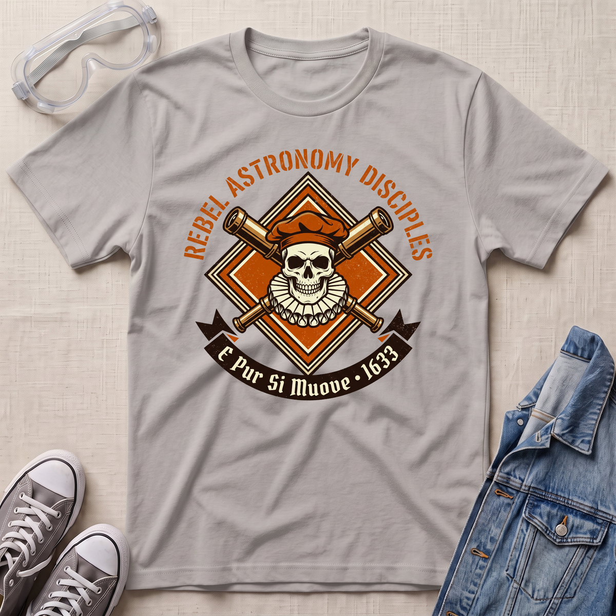 A light gray crewneck t-shirt laid flat on a textured beige surface, featuring a retro graphic of a skull wearing an orange beret with crossed telescopes and the text 'REBEL ASTRONOMY DISCIPLES' and 'E Pur Si Muove - 1633'. The shirt is styled with a pair of clear safety goggles, gray sneakers, and a blue denim jacket.