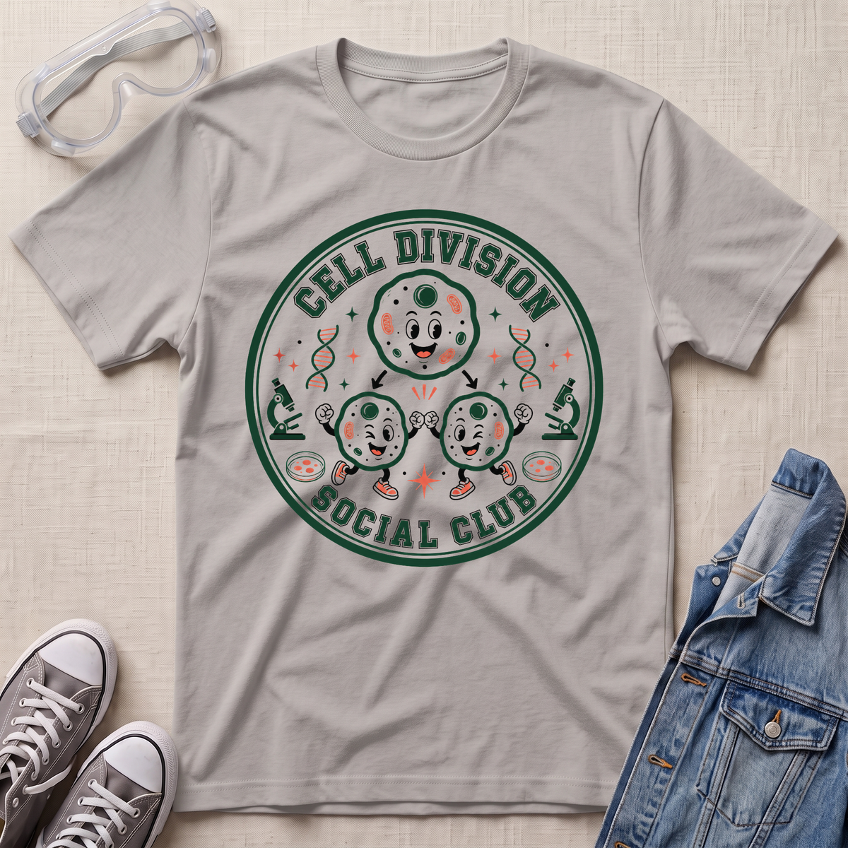 A light grey graphic t-shirt featuring a circular 'Cell Division Social Club' design with cartoon cells and microscopes, styled in a flat lay with safety goggles, sneakers, and a denim jacket on a textured beige background.
