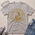 A ice gray short-sleeve graphic T-shirt laid flat on a light beige textured surface, surrounded by styling props including clear safety goggles, gray sneakers, and a blue denim jacket. The shirt features a large circular tan/gold print with a portrait and the text 'E PUR SI MUOVE CLUB AND YET IT MOVES'.