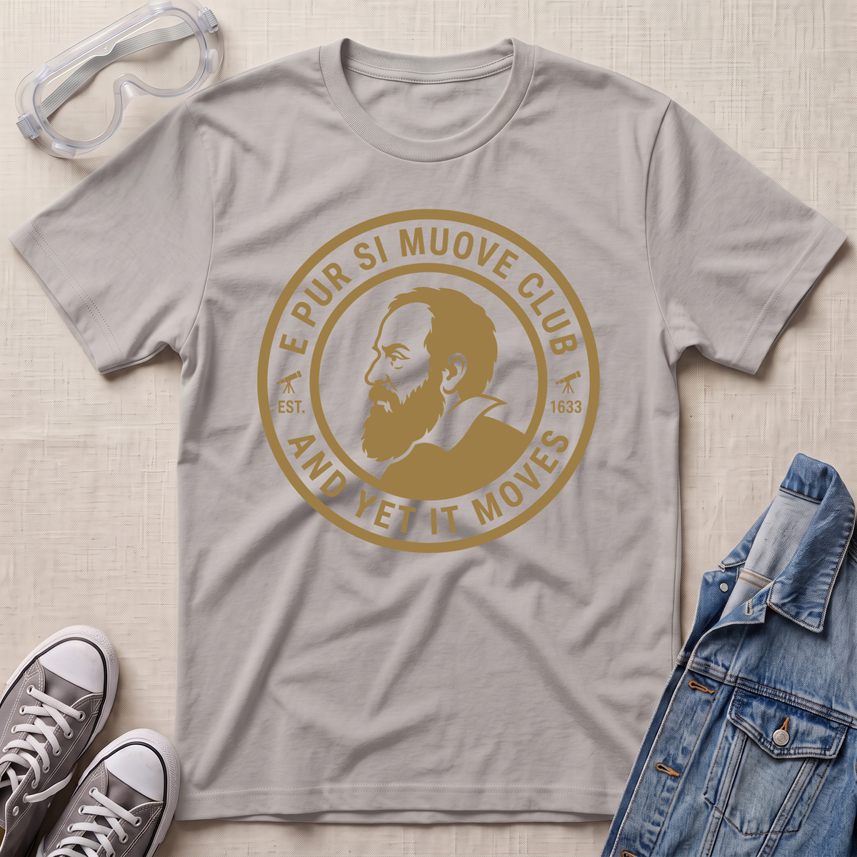 A ice gray short-sleeve graphic T-shirt laid flat on a light beige textured surface, surrounded by styling props including clear safety goggles, gray sneakers, and a blue denim jacket. The shirt features a large circular tan/gold print with a portrait and the text 'E PUR SI MUOVE CLUB AND YET IT MOVES'.