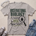 A light gray graphic t-shirt featuring the text 'OBSERVATIONAL BIOLOGY: I found a BUG. Now what?' in dark green and black, styled in a flat-lay with safety goggles, gray sneakers, and a denim jacket on a beige fabric background.