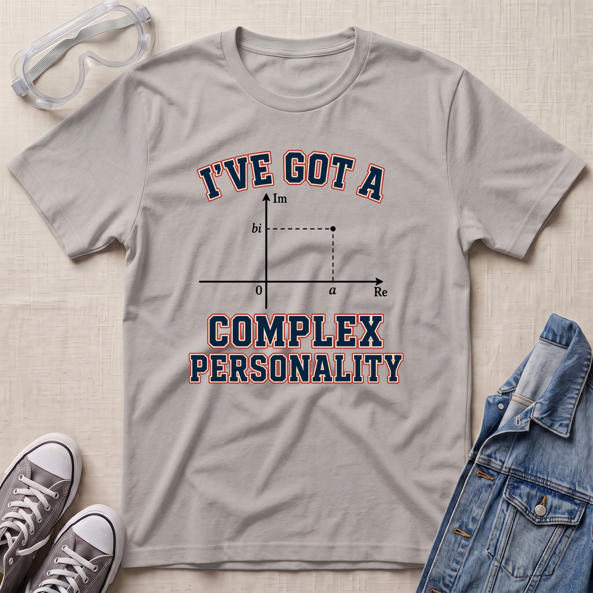 A light gray graphic t-shirt laid flat on a textured beige surface, styled with safety goggles, gray sneakers, and a blue denim jacket. The t-shirt features a graphic of a complex number plot with the text 'I'VE GOT A COMPLEX PERSONALITY' in navy blue with a red outline.