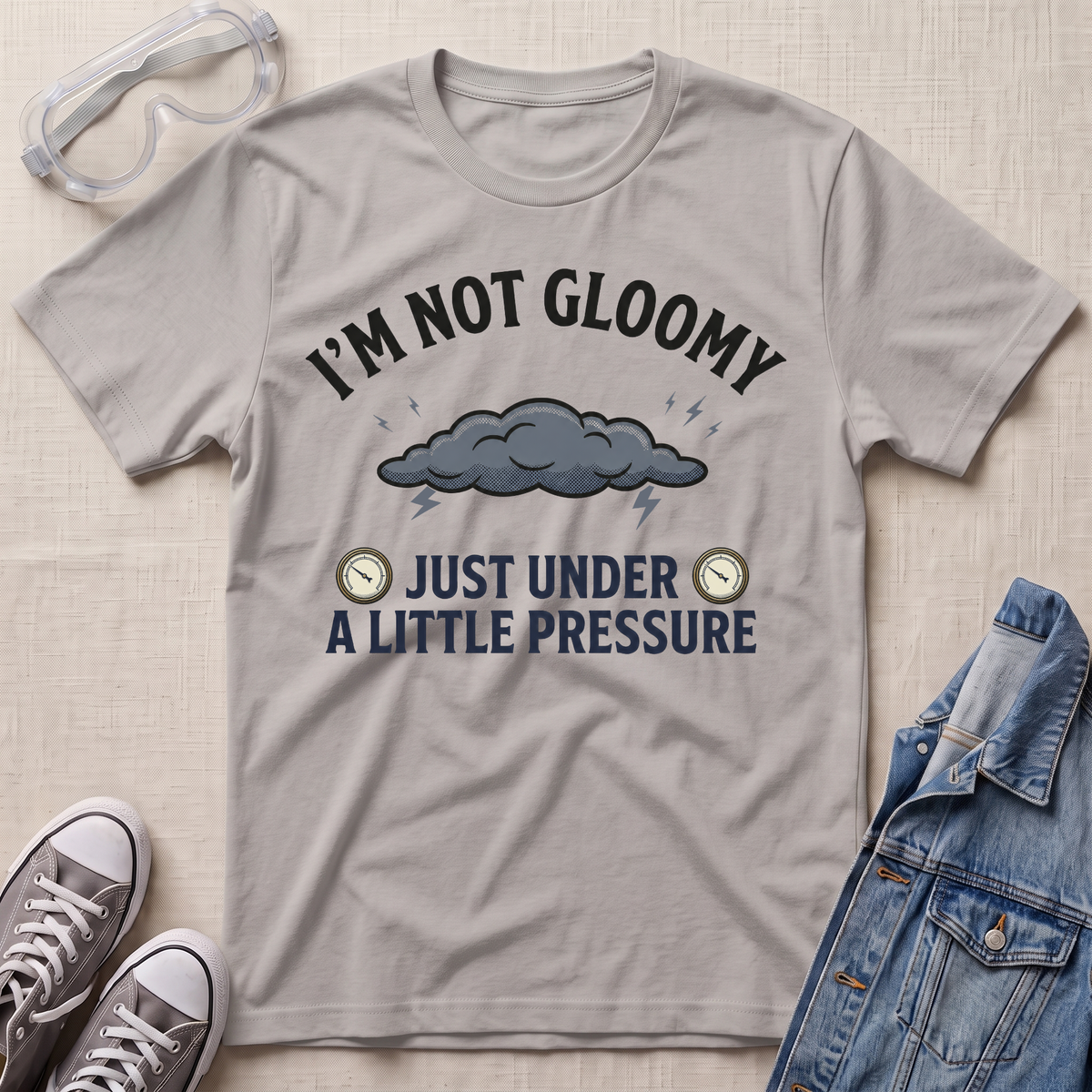 A light gray graphic t-shirt laid flat on a beige textured background, styled with safety goggles, gray sneakers, and a blue denim jacket. The t-shirt features a dark gray cloud graphic with lightning and the text 'I'M NOT GLOOMY JUST UNDER A LITTLE PRESSURE'.