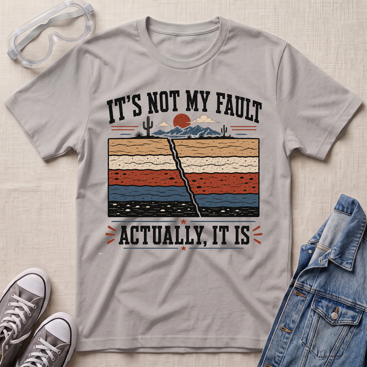 A light gray graphic t-shirt laid flat on a textured beige fabric background, styled with safety glasses, gray sneakers, and a blue denim jacket. The t-shirt features a humorous graphic with the text 'IT'S NOT MY FAULT ACTUALLY, IT IS' surrounding a retro cross-section illustration of a desert landscape.
