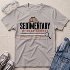 A light gray graphic t-shirt featuring a detective themed 'Sedimentary My Dear Rockson' print, styled in a flat-lay with a denim jacket, sneakers, and safety goggles on a textured beige background.