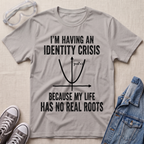 A light gray graphic t-shirt laid flat on a beige fabric background, styled with safety goggles, gray sneakers, and a blue denim jacket. The t-shirt features a black printed graphic of a parabola with the text 'I'M HAVING AN IDENTITY CRISIS BECAUSE MY LIFE HAS NO REAL ROOTS'.