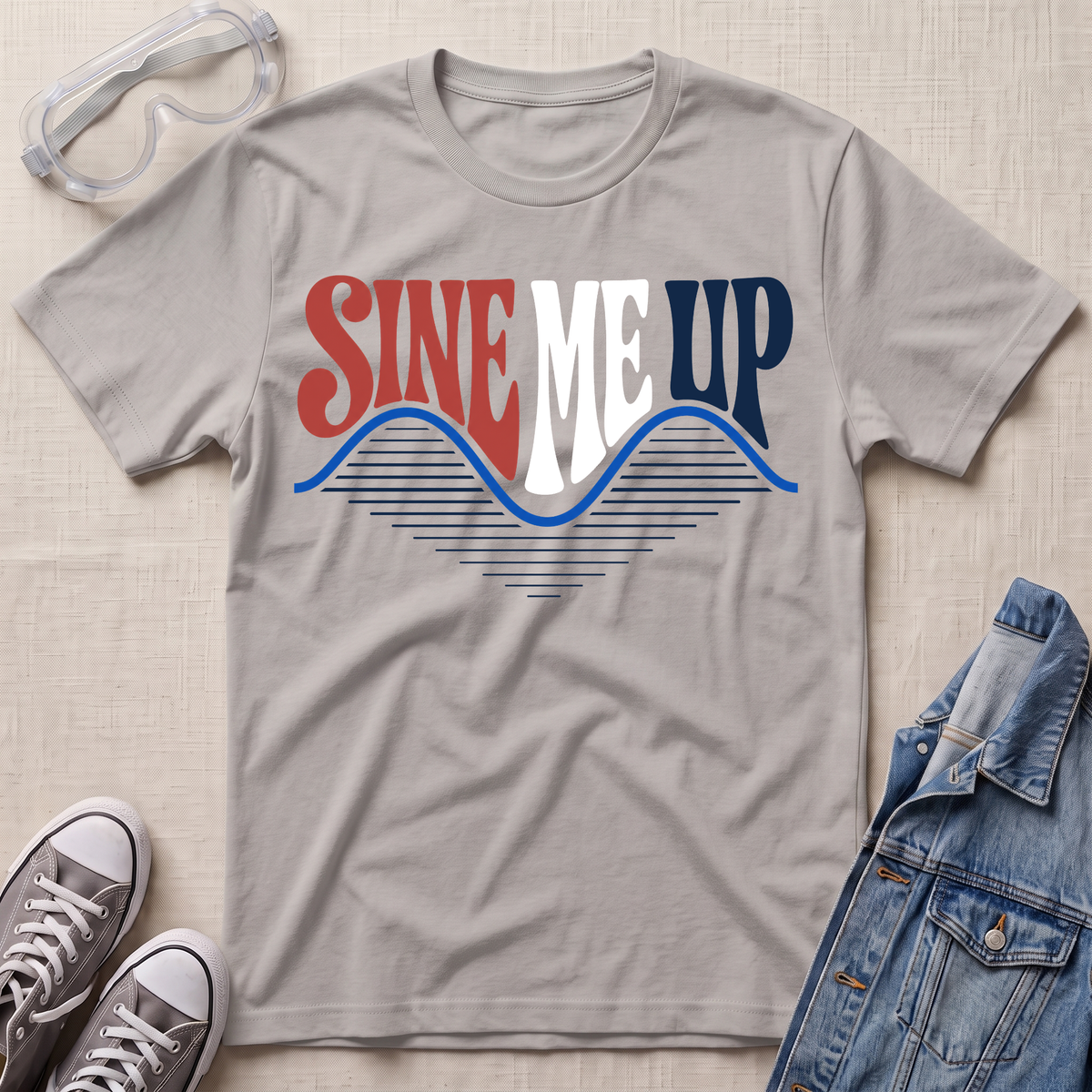A light gray t-shirt laid flat on a textured beige background, featuring a retro graphic that reads 'SINE ME UP' in red, white, and blue, styled with safety glasses, gray sneakers, and a denim jacket.