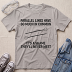 A light gray graphic t-shirt laid flat on a beige textured background, styled with safety goggles, gray sneakers, and a blue denim jacket. The t-shirt features a black printed graphic of two parallel lines with sad faces and the text 'PARALLEL LINES HAVE SO MUCH IN COMMON IT'S A SHAME THEY'LL NEVER MEET'.