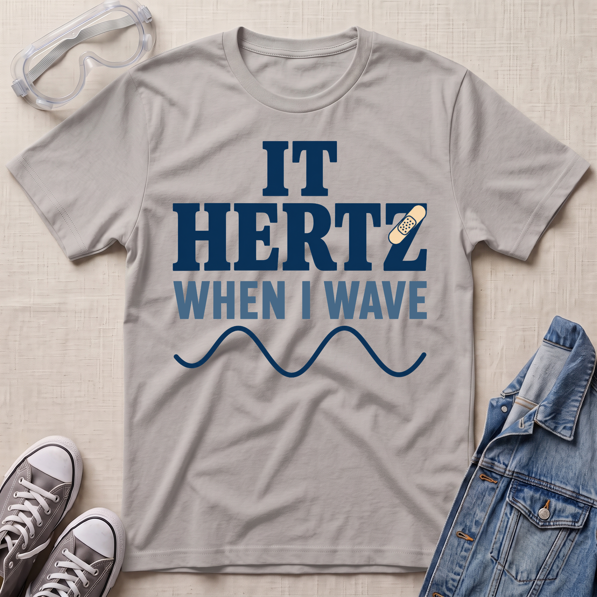 A light gray graphic t-shirt laid flat on a textured beige surface, surrounded by styling props including safety goggles, gray sneakers, and a denim jacket. The t-shirt features a dark blue and light blue graphic that reads 'IT HERTZ WHEN I WAVE' with a wavy line and a small band-aid graphic.