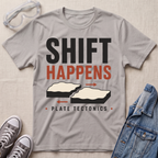 A light gray graphic t-shirt laid flat on a textured beige background, styled with safety goggles, gray sneakers, and a denim jacket. The t-shirt features a graphic that reads 'SHIFT HAPPENS' in black and orange, with illustrations of shifting tectonic plates and 'PLATE TECTONICS' underneath.