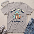 A light gray graphic t-shirt featuring a platypus wearing glasses under a blue bell curve with the text 'I'M NOT WEIRD' and 'I'M WITHIN ±1σ'. The t-shirt is styled in a flat-lay arrangement on a textured beige background, surrounded by props including safety goggles, gray sneakers, and a denim jacket.