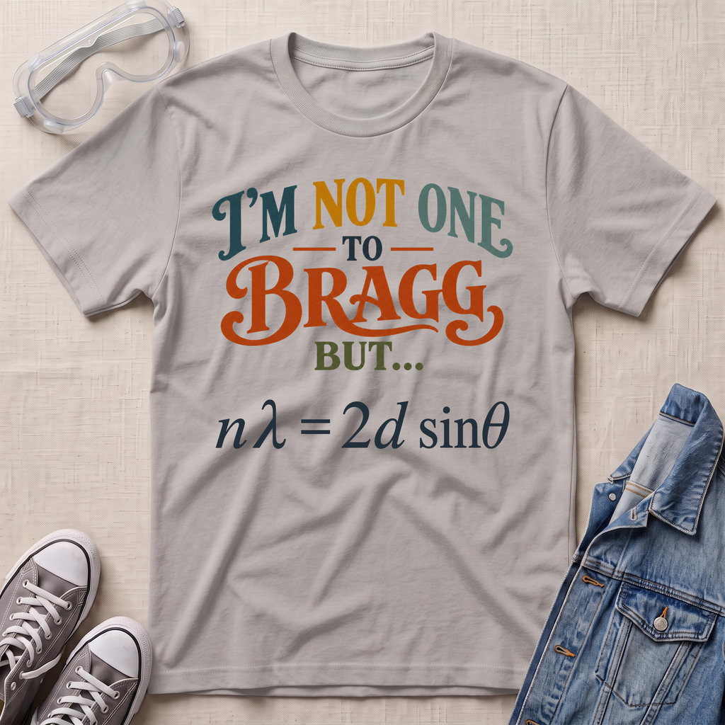 A light gray graphic t-shirt laid flat on a beige textured background, surrounded by styling props including safety goggles, gray sneakers, and a blue denim jacket. The t-shirt features a humorous physics joke with the text 'I'M NOT ONE TO BRAGG BUT.