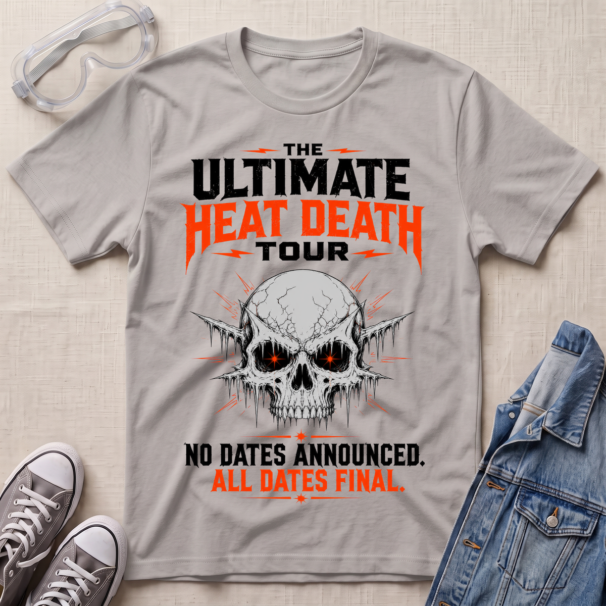 A light gray graphic t-shirt laid flat on a textured beige background, styled with safety goggles, gray sneakers, and a denim jacket. The t-shirt features a skull graphic with glowing red eyes and bold text that reads 'THE ULTIMATE HEAT DEATH TOUR' and 'NO DATES ANNOUNCED.