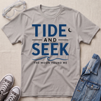 A ice gray short-sleeve t-shirt with a blue graphic reading 'TIDE AND SEEK THE MOON FOUND ME' laid flat on a beige textured surface, surrounded by styling props including safety goggles, gray sneakers, and a denim jacket.