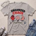 A light gray graphic t-shirt laid flat on a beige textured background, styled with safety goggles, gray sneakers, and a denim jacket. The t-shirt features a cartoon graphic of a heart, a brain, and a stomach with the text 'ORGANIZED CHAOS' and 'HUMAN BODY DEPT.