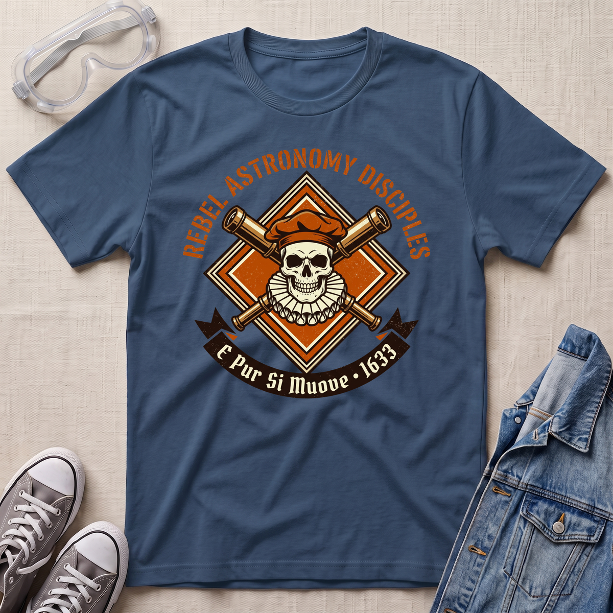 A indigo blue graphic t-shirt laid flat on a light beige fabric background, styled with clear safety goggles, gray sneakers, and a denim jacket. The t-shirt features a central graphic of a skull wearing an orange beret, with crossed telescopes behind it, and the text 'REBELLION ASTRONOMY DISCIPLINE' and 'E Pur Si Muove - 1633'.
