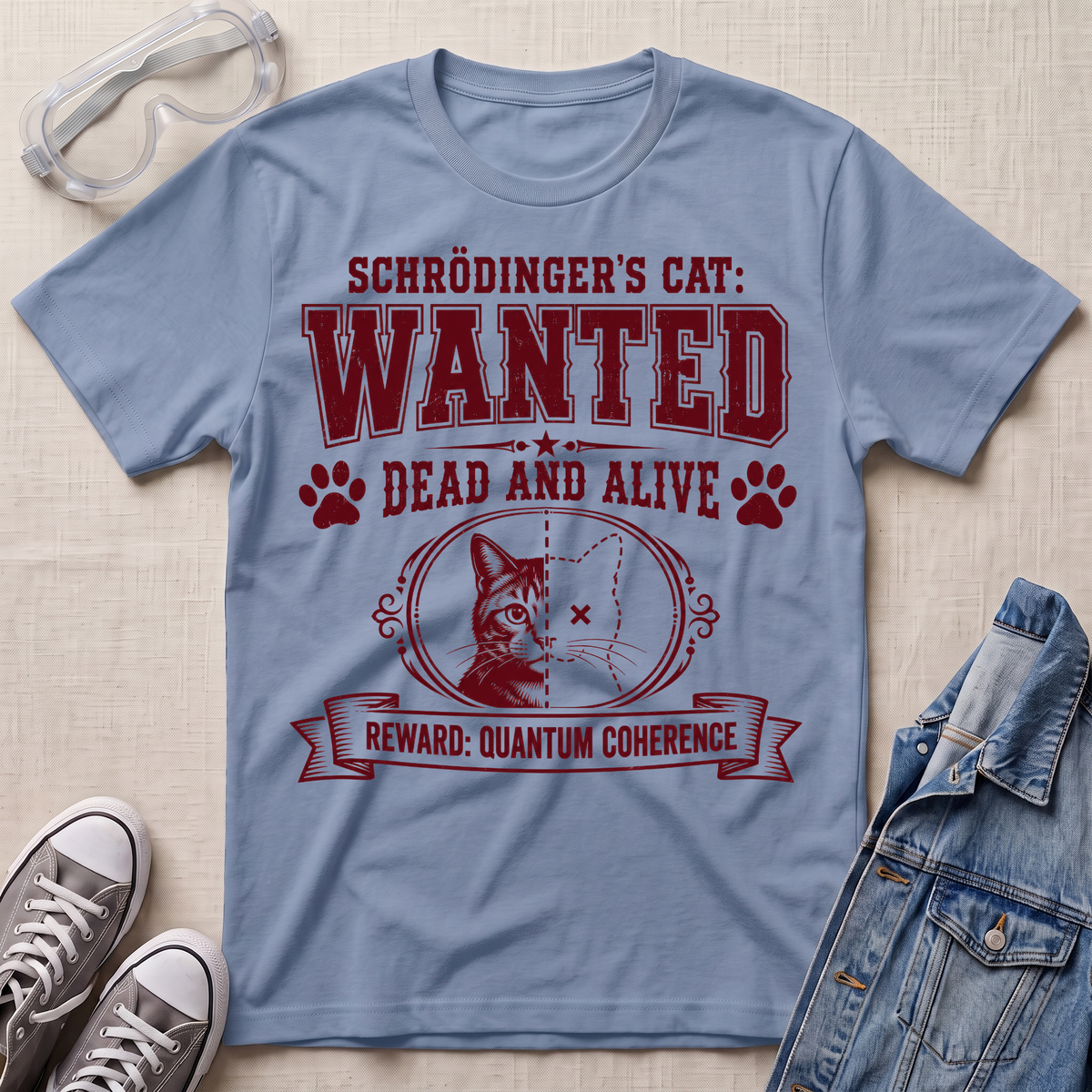 A light blue graphic t-shirt laid flat on a beige fabric background, styled with safety goggles, gray sneakers, and a denim jacket. The t-shirt features a maroon graphic of Schrödinger's cat with the text 'SCHRÖDINGER'S CAT: WANTED DEAD AND ALIVE REWARD: QUANTUM COHERENCE'.