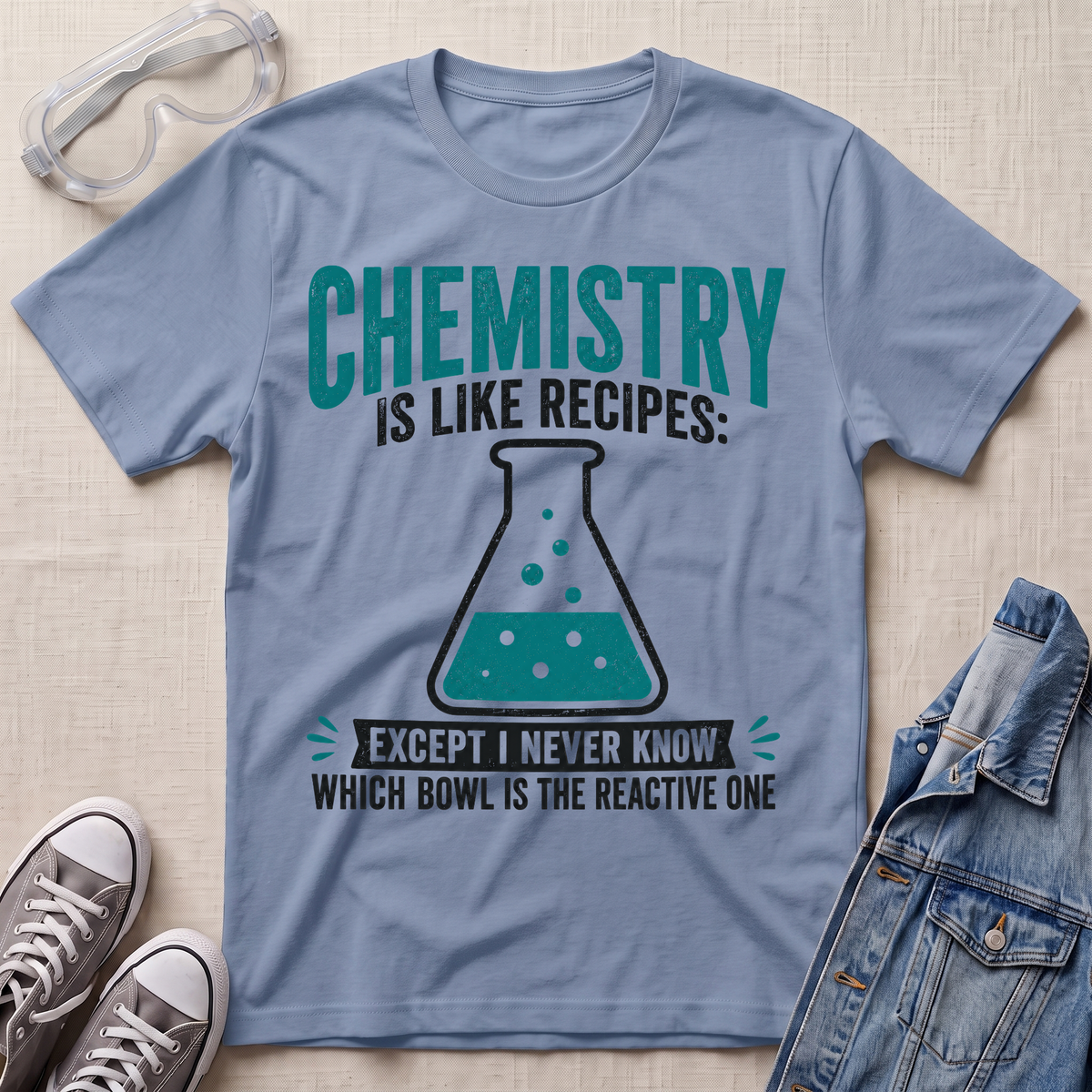 A light blue graphic t-shirt laid flat on a beige fabric background, styled with safety goggles, gray sneakers, and a denim jacket. The t-shirt features a teal and black graphic that reads 'CHEMISTRY IS LIKE RECIPES: EXCEPT I NEVER KNOW WHICH BOWL IS THE REACTIVE ONE' with an illustration of a beaker.
