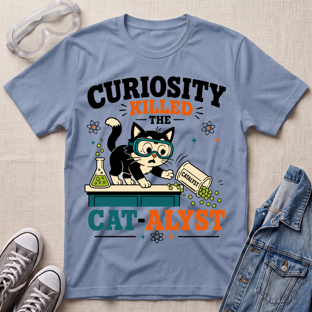 A light blue graphic T-shirt laid flat on a textured beige surface, surrounded by styling props including safety goggles, gray sneakers, and a denim jacket. The shirt features a cartoon cat wearing goggles and pushing a spilled pill bottle labeled 'CAT-ALYST' under the words 'CURIOSITY KILLED THE CAT-ALYST'.